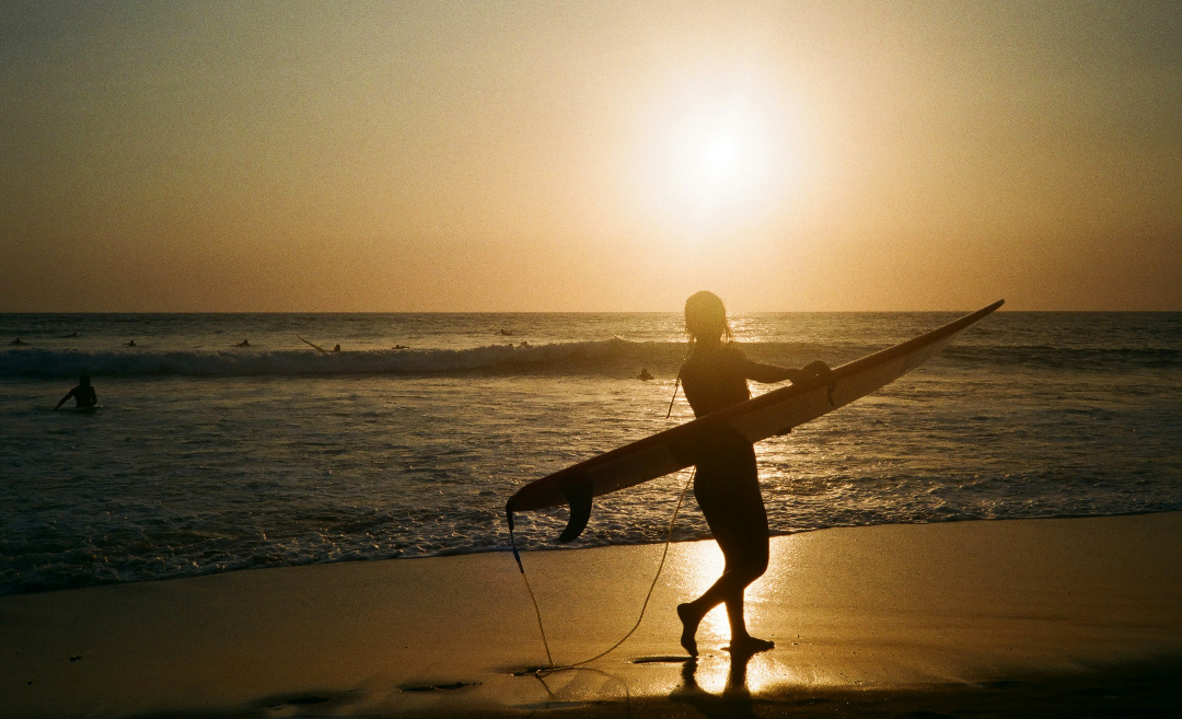 Silhouette of a woman standing on the beach holding a surfboard at sunset in Canggu, Bali.