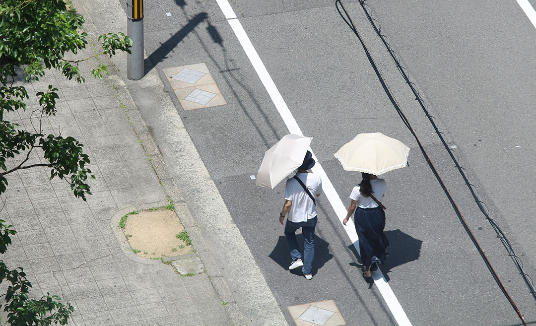 Pedestrians walking along a sunny city street holding umbrellas to shelter from bright sunlight