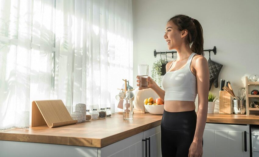 Woman in activewear drinking water from a bottle inside her home after a workout