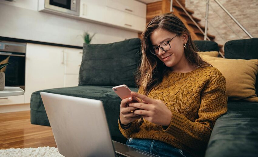 Woman working on a laptop and checking her smartphone while sitting comfortably on the floor at home.