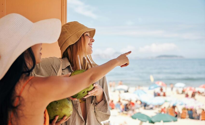 Two women with coconut drinks pointing at a crowded beach, illustrating the choice of slow travel over overcrowded tourist spots.