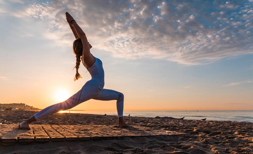 Woman practicing yoga on a beach at sunrise to start a gentle morning of travel.