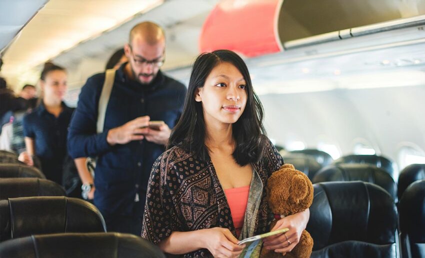 Female traveller in airplane aisle with carry-on items waiting to deboard, illustrating passenger vulnerability.