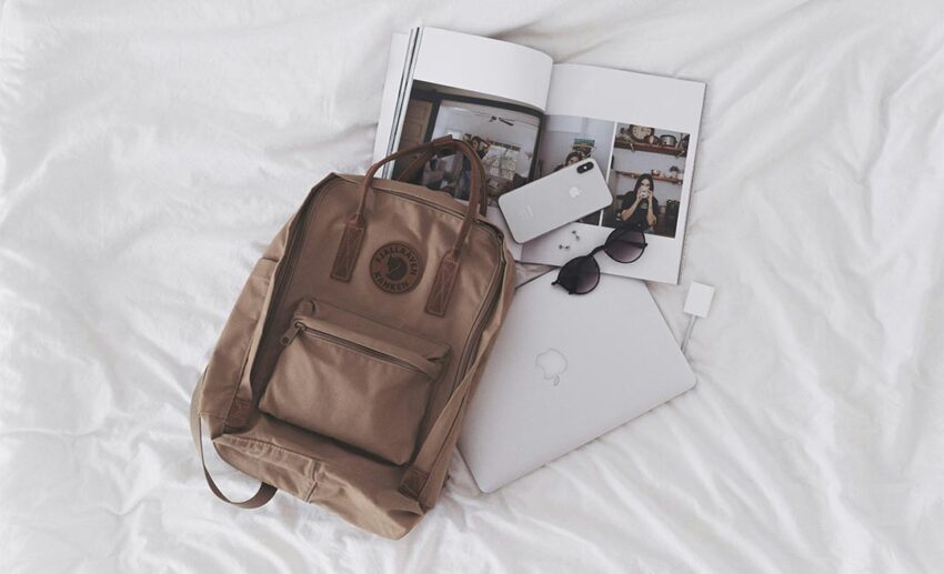 Travel flat lay with a brown backpack, laptop, iPhone, and sunglasses on a white background.