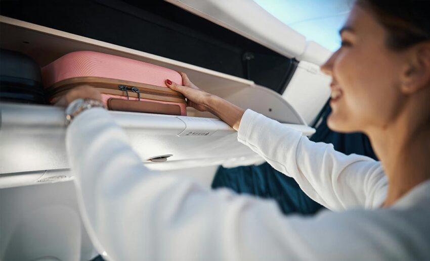 Smiling woman in white shirt stowing a pink hard-shell carry-on suitcase in an airplane overhead bin.