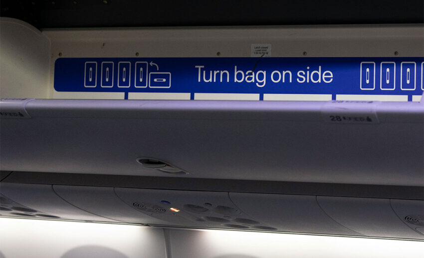 Open airplane overhead storage bin with instructions to turn bag on side, showing shared cabin space.