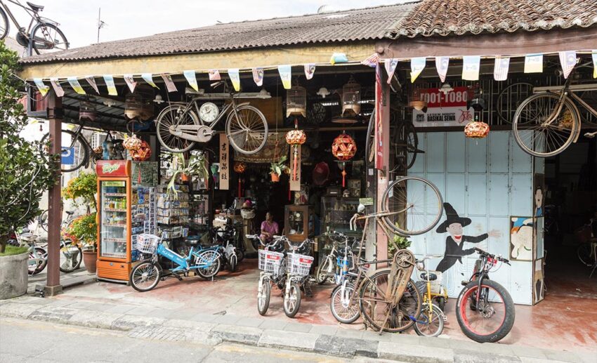 A traditional Malaysian sundry shop with a wooden storefront, vintage bicycles, a glass drink fridge, and miscellaneous household items for sale.