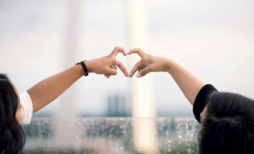 Two female friends standing on a scenic overlook with their arms raised and fingers touching to form a heart shape.
