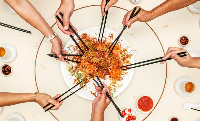 Overhead shot of hands using chopsticks to participate in the Lou Sang or Yee Sang prosperity toss, a raw fish salad tradition in Malaysia and Singapore.