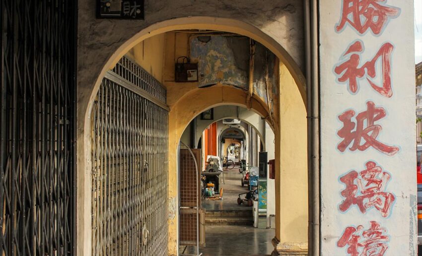 A perspective shot of a long corridor walkway outside a row of heritage pre-war shophouses with weathered pillars and shadows.