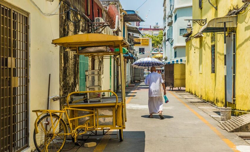 A back view of an elderly man walking down a quiet heritage backlane in Malaysia, holding an umbrella for shade.