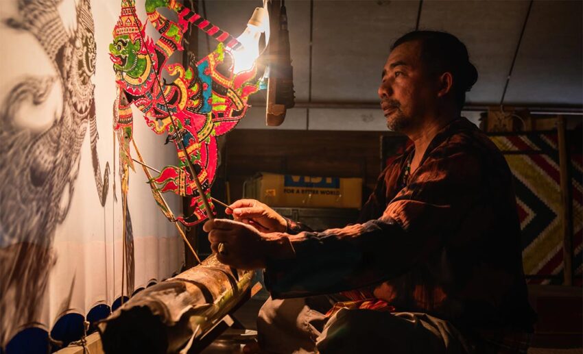 A view from behind the shadow screen showing a Tok Dalang (master puppeteer) manipulating leather puppets during a traditional Wayang Kulit performance.