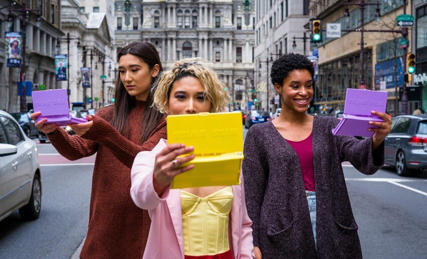 Three women standing on a city street, huddled together and looking into a makeup palette mirror.