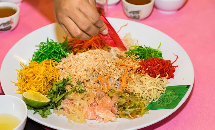 A close-up shot of a person’s arm pouring a packet of toasted sesame seeds onto a plate of Yee Sang during a Chinese New Year celebration.