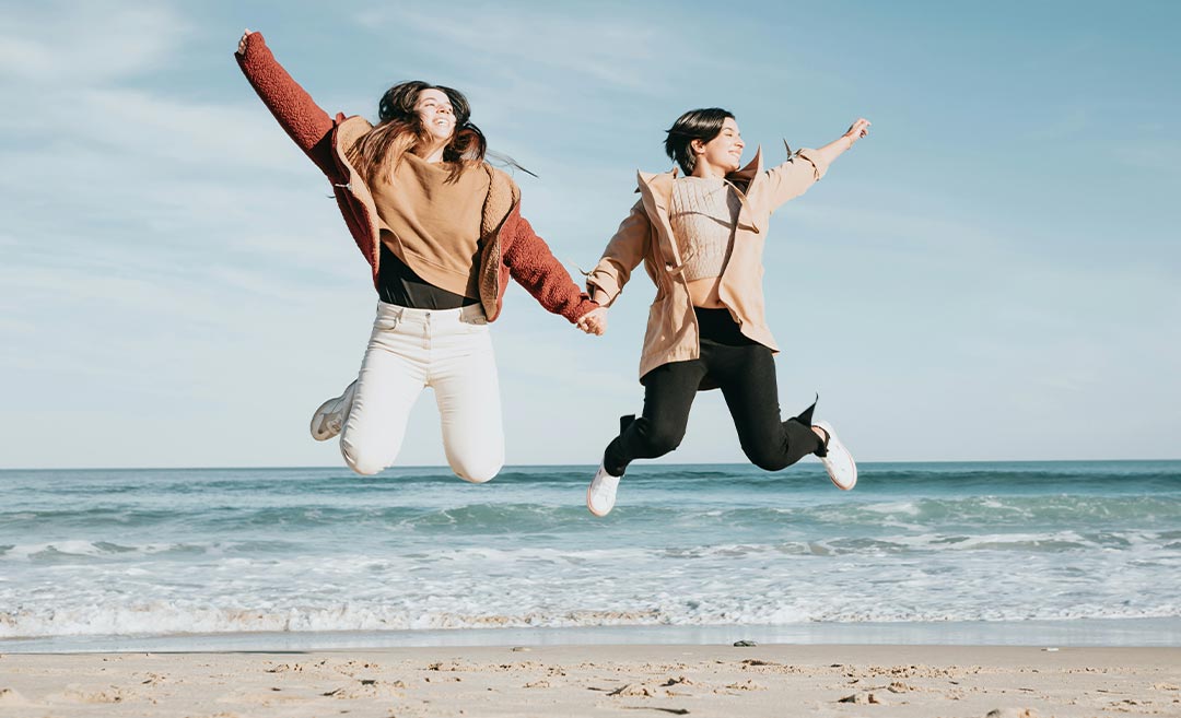 Two women holding hands and jumping gleefully on a sun-drenched beach during a girls' trip.