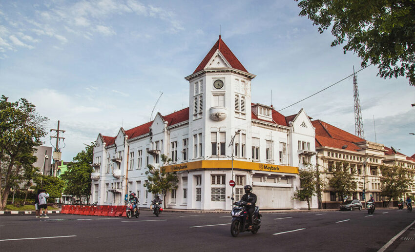 A man riding a motorcycle past a historic colonial-style bank building on a street in Surabaya, Indonesia.