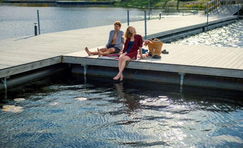A man and woman relaxing and lounging on a wooden pier dock over a calm Finnish lake in the summer.