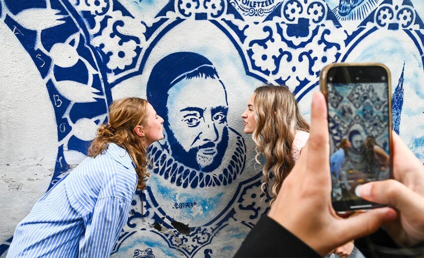Two women posing in front of a large-scale blue and white ceramic-inspired mural on a brick wall, blending Delft’s historic pottery heritage with modern street art.