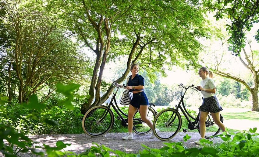 Two women walking their bicycles through a shaded, leafy path in Delft, illustrating the town’s flat landscape and unhurried cycling culture.