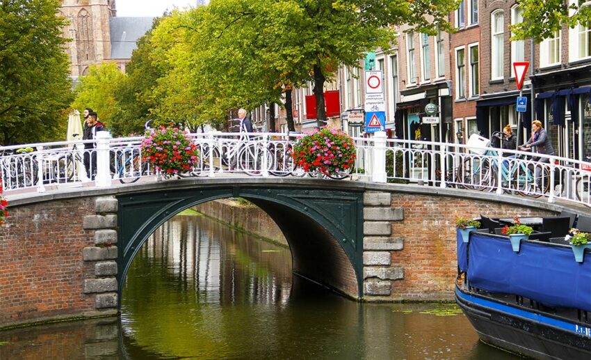 Pedestrians strolling across a historic bridge with white railings over a serene canal in Delft, showcasing a quieter, crowd-free alternative to Amsterdam.