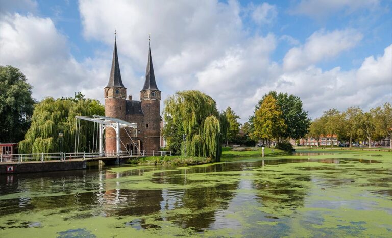 The historic Oostpoort or East Gate in Delft, featuring 15th-century twin towers and a wooden drawbridge over a calm canal, reflecting the soft Golden Age light that inspired Vermeer’s paintings.