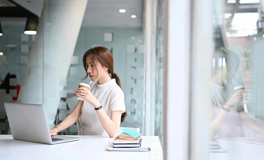 A woman sitting at a modern workplace near a window, holding a coffee cup while checking flight updates on her laptop.