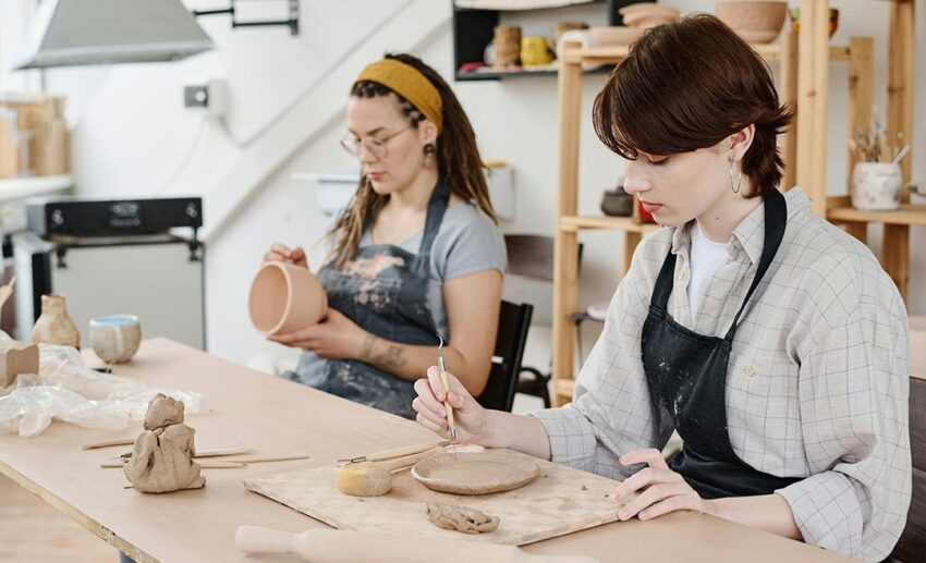 Two women wearing aprons sitting at a table together, using tools to cut and shape clay ornaments for a pottery project.