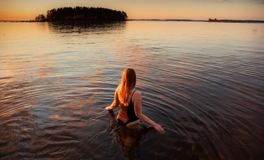 A woman in a bathing suit standing waist-deep in the clear water of a lake in Kotka, Finland, preparing for a swim.