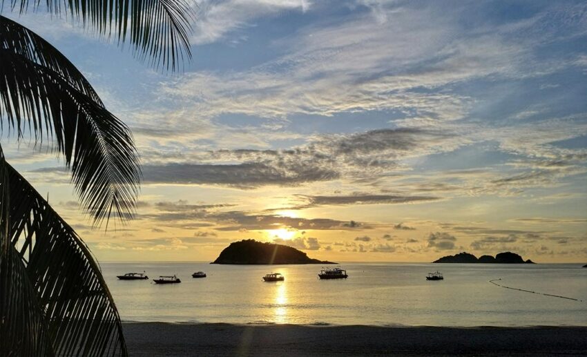 A scenic sunrise view from a beach in Kuala Terengganu showing fishing boats anchored in the water and small islands on the horizon.