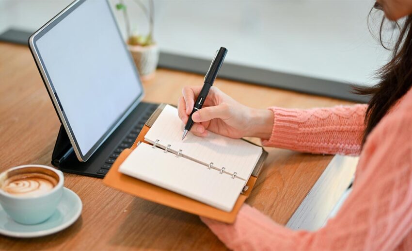 A woman using a digital tablet and a paper personal planner book to schedule her weekly creative goals and artistic projects.