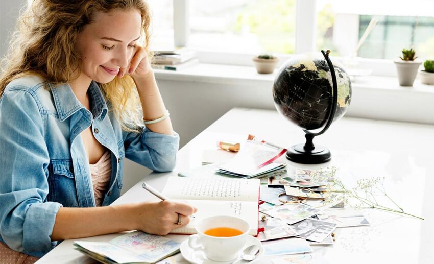 A woman writing in a travel diary surrounded by photo prints, a passport, and a tabletop globe, planning a long-duration creative getaway.