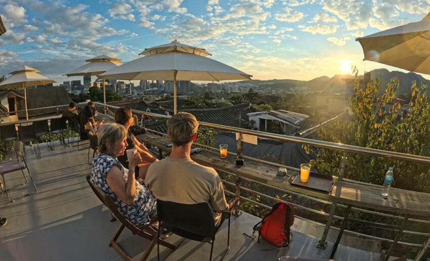 People sitting at an outdoor rooftop cafe in Bukchon Hanok Village overlooking traditional Korean houses and distant mountains.