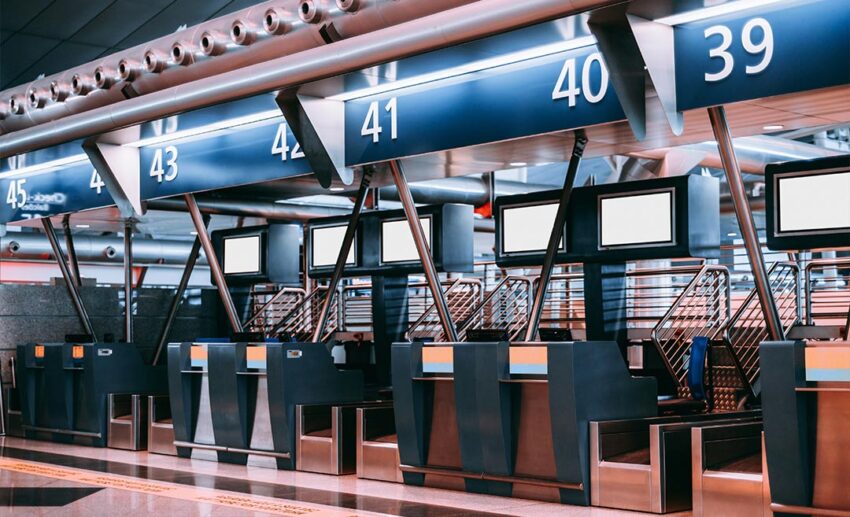 The interior of a modern airport check-in area featuring automated baggage handling systems and blank white information screens.