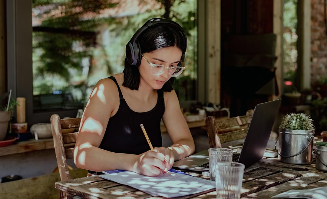 A woman sitting at a table wearing headphones, working on a laptop and writing in a notebook during a structured creative retreat.