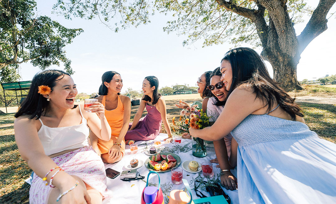 A group of women celebrating International Women's Day 2026 with a colourful picnic in a park in Kuala Lumpur, featuring floral decorations and fresh fruits.