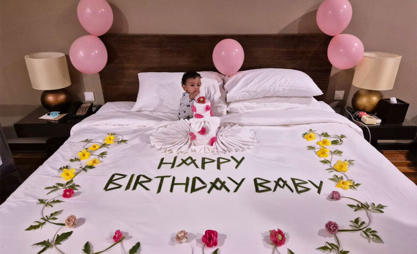 A one-year-old baby sitting happily on a resort bed decorated by staff with green leaves spelling out "Happy Birthday Baby," surrounded by flowers and a towel folded like a birthday cake.