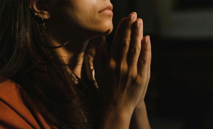 A woman in an orange shirt with her hands pressed together in a prayer-like gesture, symbolising a universal sense of the sacred.