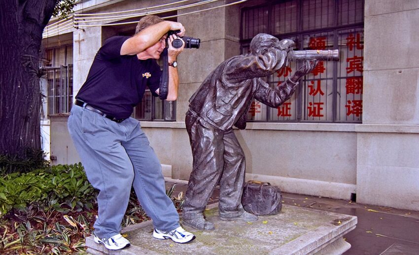 A young David Bowden posing with a long-lens camera next to a statue of a photographer in a similar pose.