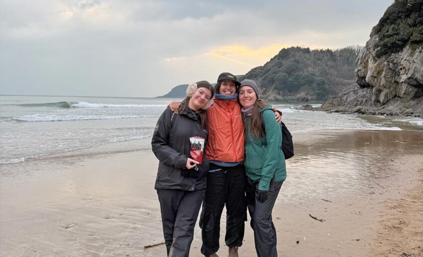 Sofia and two female friends in a group hug on the sandy beach at Caswell Bay, South Wales.