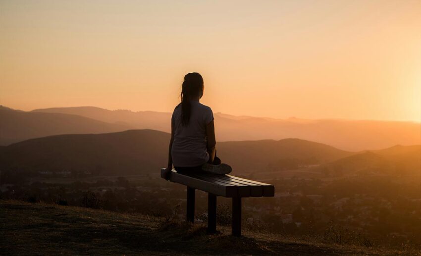 A woman sitting on a wooden bench overlooking a vast, misty mountain range, reflecting quietly during a holiday away from home.