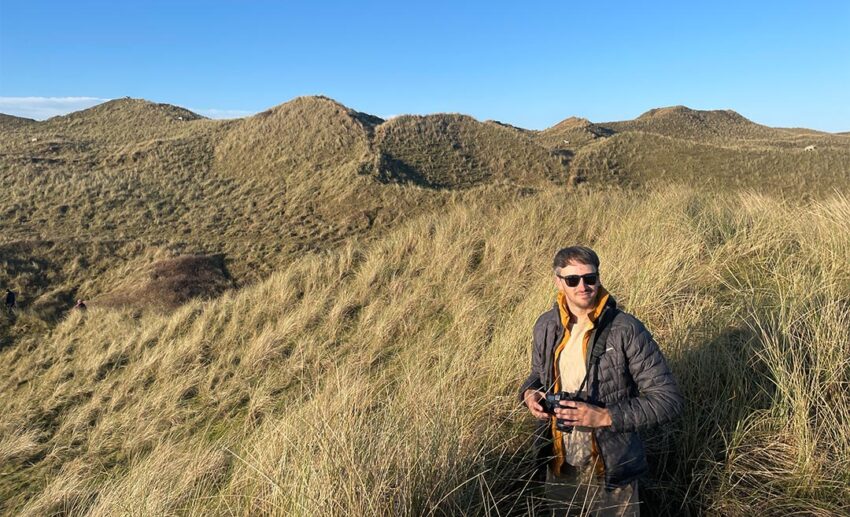 A man standing on the grassy cliffs of Rhossili Bay, looking out over the dramatic South Wales coastline.