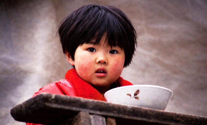 A close-up of a young Asian child in a red jacket sitting in front of a white bowl, looking directly at the camera.