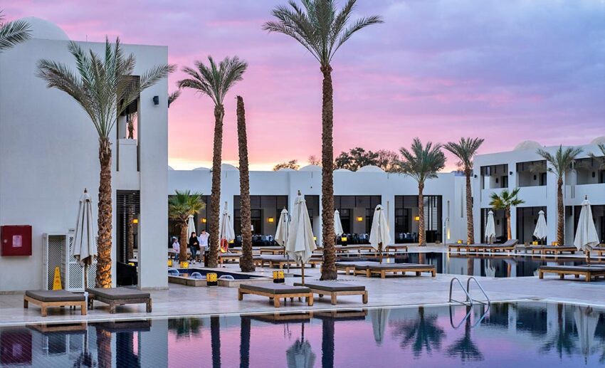ground-floor view of a luxury swimming pool surrounded by palm trees at sunset.