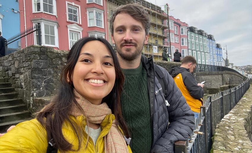 Sofia and her partner taking a selfie in front of a row of iconic pastel-coloured harbour buildings in Tenby, Wales.