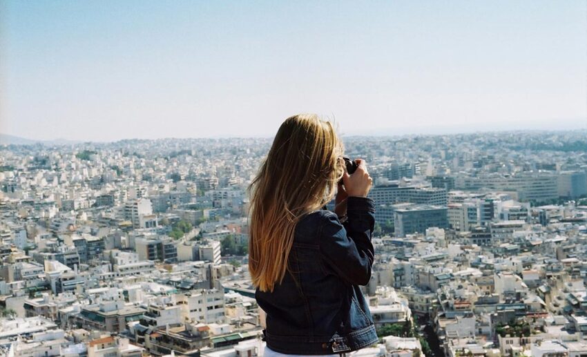 A woman standing at a high viewpoint in Athens, Greece, taking a photograph of the sprawling city landscape below.