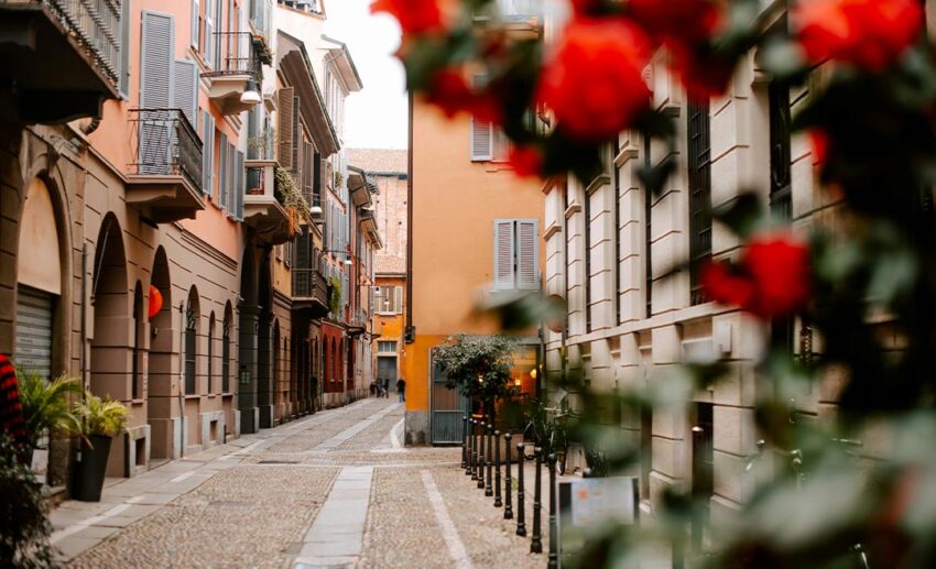 A narrow, traditional street in Milan, Italy, featuring balconied buildings decorated with flowering plants.
