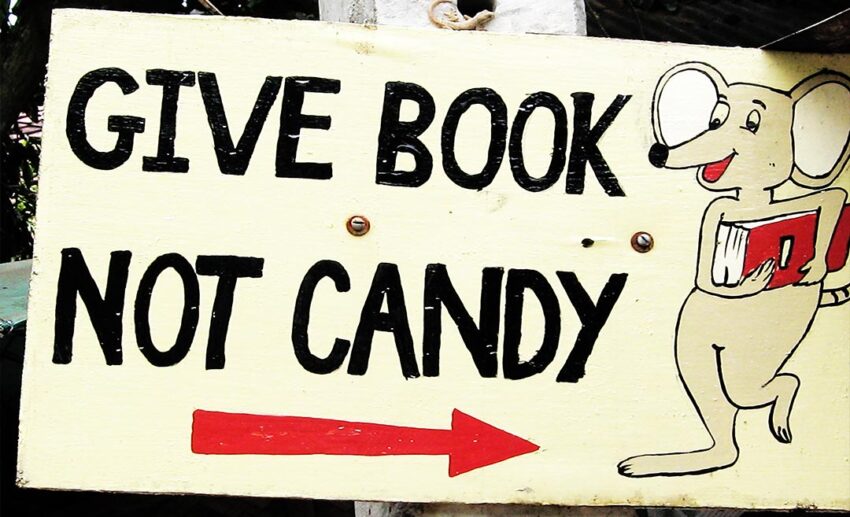 A hand-painted sign featuring a mouse holding a book with the text "Give Book, Not Candy," encouraging ethical gifting in tourism.
