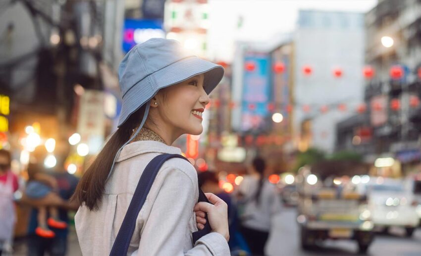 A side profile of a smiling woman wearing a bucket hat and a sling bag, walking along a vibrant, busy street in Bangkok, Thailand.