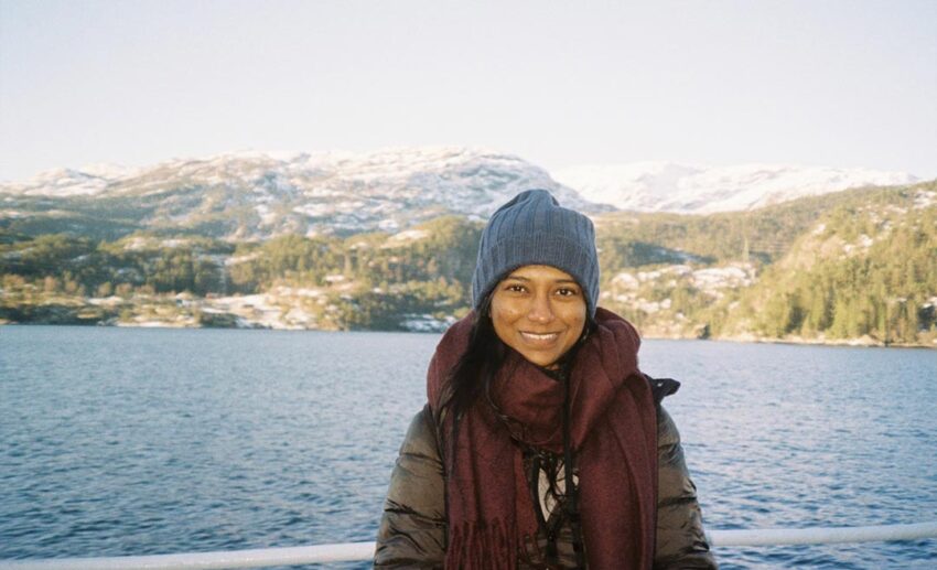 A female traveller, Tabitha, smiling on the deck of a fjord cruise boat with the steep, snow-dusted hills and mountains of Bergen in the background.