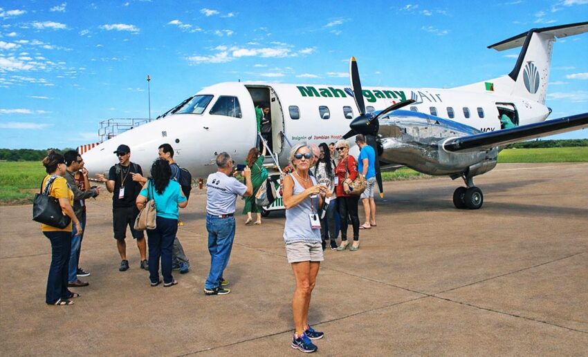 Passengers standing near a Mahogany Air propeller plane on the tarmac of a small airport in Zambia.